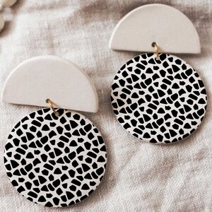 Puede incluir: Un par de pendientes de cerámica blanca con círculos de patrones geométricos en blanco y negro. Los pendientes tienen forma de semicírculos con un pequeño bucle dorado en la parte superior.
