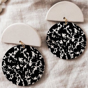 Puede incluir: Un par de pendientes con estampado en blanco y negro con detalles en forma de semicírculo blanco. Los pendientes están hechos de arcilla y tienen un diseño moderno y abstracto.