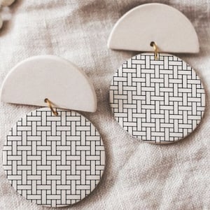 Pode incluir: Um par de brincos de cerâmica branca com um padrão geométrico. Cada brinco apresenta uma forma de semicírculo sobre uma forma redonda com um design tecido em preto e branco.