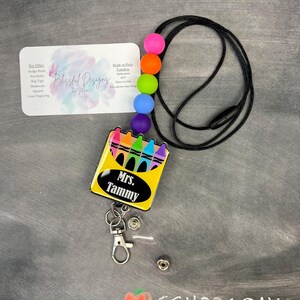 K&ouml;nnte beinhalten: Ein schwarzes Lanyard mit bunten Silikonperlen und einem einziehbaren Ausweishalter. Der Ausweishalter zeigt eine gelbe Box mit einer Reihe von Buntstiften und dem Text "Mrs. Tammy".