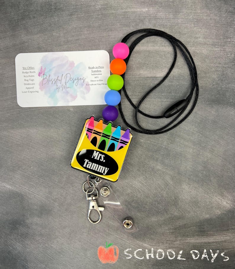 K&ouml;nnte beinhalten: Ein schwarzes Lanyard mit bunten Silikonperlen und einem einziehbaren Ausweishalter. Der Ausweishalter zeigt eine gelbe Box mit einer Reihe von Buntstiften und dem Text "Mrs. Tammy".