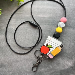 Könnte beinhalten: Ein schwarzes Schlüsselband mit einem Schul-Themen-Anhänger. Der Anhänger zeigt einen roten Apfel, einen gelben Bleistift und einen Notizblock. Das Schlüsselband hat auch bunte Perlen und einen Metallverschluss. Die Worte "SCHOOL DAYS" stehen unten.