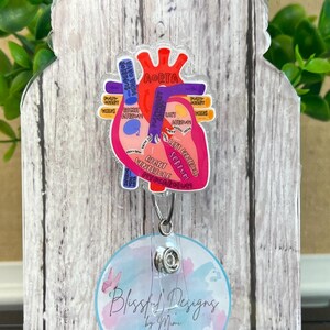 Könnte beinhalten: Ein transparenter, herzförmiger Badgehalter aus Acryl mit einer farbenfrohen anatomischen Herzillustration. Die Illustration enthält Beschriftungen für die Aorta, die obere Hohlvene, den rechten Vorhof, die rechte Kammer, den linken Vorhof, die linke Kammer und das Septum. Der Badgehalter ist an einer silbernen, einziehbaren Badge-Rolle mit einem transparenten Plastikband befestigt. Der Badgehalter befindet sich auf einem Holzgrund mit einem weißen und blauen Aquarelldesign und dem Text "Blissful Designs by Mimi".
