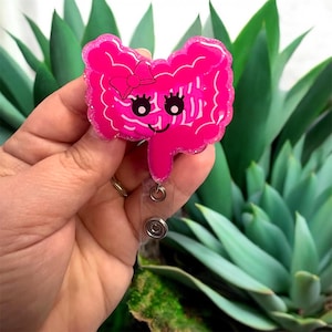 Könnte beinhalten: Ein pinkfarbenes, glitzerndes Ausweis-Jojo in Form eines Cartoon-Dickdarms mit einem lächelnden Gesicht und einer Schleife. Das Ausweis-Jojo hat eine transparente, ausziehbare Kunststoffkordel und einen Metallclip.