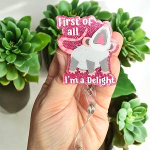 Könnte beinhalten: Rosa glitzernder Badgehalter mit einem Opossum-Cartoon und dem Text "First of all I'm a Delight".
