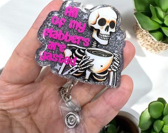 Peut inclure: Un porte-badge avec une bordure grise pailletée et un motif de squelette. Le squelette tient une tasse de café et le texte dit "All of my stabbers are fasted" en lettres roses. Le porte-badge a une sangle en plastique transparent et un clip en métal.