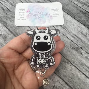 Könnte beinhalten: Ein einziehbares Ausweishalter mit einer silbernen Glitzerkuh mit Skelett-Design. Die Kuh hat schwarze Augen und eine schwarze Nase. Der Ausweishalter ist aus klarem Kunststoff.