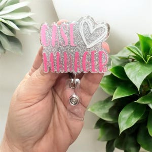 Könnte beinhalten: Ein glitzerndes Ausweis-Jojo mit den Worten "CASE MANAGER" in rosa Schrift. Oben rechts befindet sich ein weißes Herzdesign. Das Ausweis-Jojo hat ein durchsichtiges, einziehbares Kunststoffkabel und einen Metallclip.
