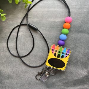 K&ouml;nnte beinhalten: Ein schwarzes Lanyard mit einem Regenbogen-Silikonperlen-Anh&auml;nger und einer gelben und schwarzen Box mit einem Regenbogen aus Buntstiften. Die Box hat ein transparentes Plastikfenster und einen schwarzen Kreis f&uuml;r ein Namensschild. Das Lanyard hat einen durchsichtigen Plastik-Badgehalter mit einem Metallclip.