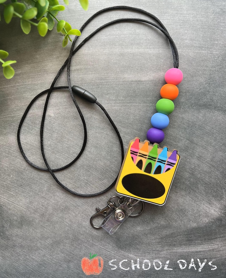 K&ouml;nnte beinhalten: Ein schwarzes Lanyard mit einem Regenbogen-Silikonperlen-Anh&auml;nger und einer gelben und schwarzen Box mit einem Regenbogen aus Buntstiften. Die Box hat ein transparentes Plastikfenster und einen schwarzen Kreis f&uuml;r ein Namensschild. Das Lanyard hat einen durchsichtigen Plastik-Badgehalter mit einem Metallclip.