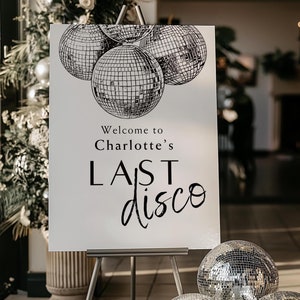以下が含まれることがあります： 「Charlotte's Last Discoへようこそ」という黒い文字が書かれた白い看板に、ミラーボールの絵が描かれている。看板はイーゼルに置かれ、その前に銀色のミラーボールが3つ置かれている。