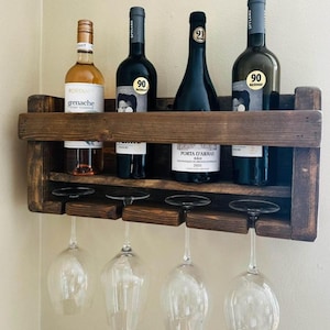 May include: A rustic wooden wine rack with four bottles of wine and four wine glasses hanging below. The rack has two shelves and is stained a dark brown color. The bottles are all different colors and have labels on them. The glasses are clear and have a stem.