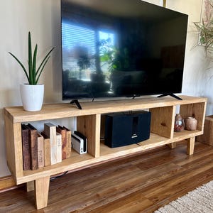 May include: A wooden TV stand with four shelves, featuring a flat screen TV, a speaker system, and books. The stand has a natural wood finish and is supported by two legs.
