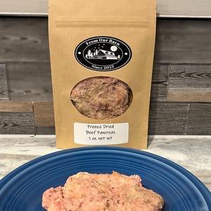 May include: A brown paper bag with a white logo that says "From Our Barn" and "Since 2023". The bag contains freeze-dried beef pancreas, with a label that says "Freeze Dried Beef Pancreas 1 oz net wt". The bag is sitting on a blue plate with a pile of freeze-dried beef pancreas.