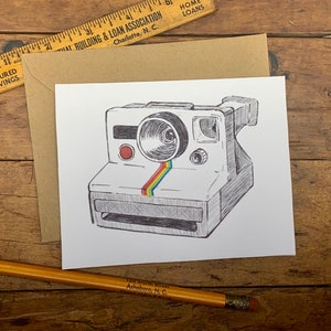 May include: A black and white line drawing of an instant camera with a rainbow stripe across the front. The camera is on a white background.