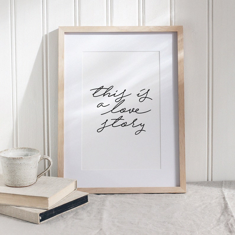 May include: A framed print with the text "this is a love story" written in black cursive script on a white background.