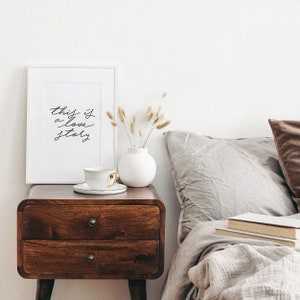 May include: A white framed print with the text "this is a love story" on a wooden nightstand with a white vase and dried flowers. A cup and saucer are on the nightstand. A bed with a gray duvet and pillows is in the background.