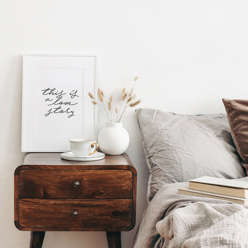 May include: A white framed print with the text "this is a love story" on a wooden nightstand with a white vase and dried flowers. A cup and saucer are on the nightstand. A bed with a gray duvet and pillows is in the background.