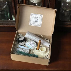 May include: A brown cardboard box with the text "The Smudge Box" and "Candle Monkey" on the lid. The box is open and contains a variety of items, including a small jar of smudge, a seashell, a wooden stick, a candle holder, and a bundle of incense sticks.