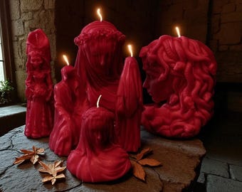 May include: A collection of red sculpted candles, each with a unique design, including figures and faces. The candles are lit, with flames visible. The candles are arranged on a stone surface, with a few decorative leaves.