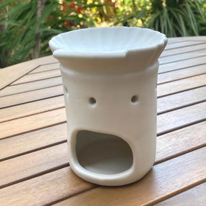 May include: A cream-colored ceramic essential oil diffuser with a wide top and a large opening at the bottom. The diffuser has three small holes and sits on a wooden surface. The design is simple and modern, suitable for home fragrance.