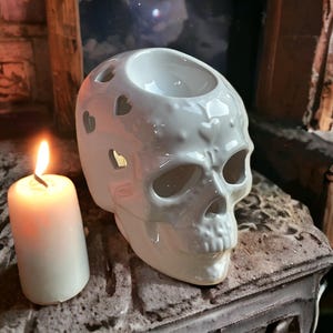 May include: White ceramic skull shaped oil burner with heart cutouts. The burner has a small well in the top for a tea light candle.