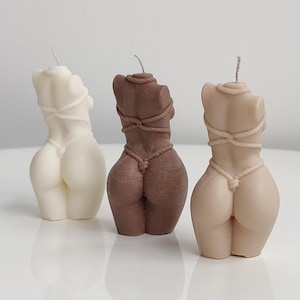 Puede incluir: Tres velas con forma de cuerpo en marfil, marrón y beige. Cada vela está esculpida para parecer un torso, con detalles de cuerda alrededor de la parte superior del cuerpo y la cintura. Las velas están diseñadas para ser decorativas y son adecuadas para la decoración del hogar.