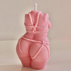 May include: Pink candle shaped like a female torso with rope detailing. The candle is tied with rope around the waist and chest.