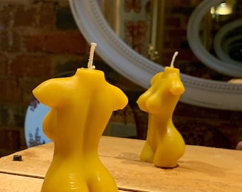 Op de afbeelding: Twee gele, lichaamsvormige kaarsen. De kaarsen hebben de vorm van een vrouwelijk lichaam, met zichtbare borsten en billen. De kaarsen staan op een houten oppervlak, met een spiegel op de achtergrond.
