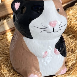 May include: A ceramic guinea pig shaped wax warmer with a black, white, and brown color scheme. The warmer has a pink nose and pink feet.