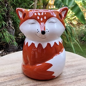 May include: A ceramic fox figurine with a smiling face and closed eyes. The fox is primarily orange with white accents on the face, chest, and tail. The figurine is sitting on a wooden surface.