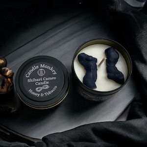 May include: A black candle tin with the text "Candle Monkey" and "Shibari Cameo Candle Honey & Tobacco". The candle is in a clear glass jar with two dark blue wax figures inside.