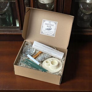 May include: A brown cardboard box with the text "The Smudge Box" on the lid. The box is open and contains a variety of items for smudging, including a white ceramic bowl, a bundle of sage, a wooden stick, a pack of incense sticks, and a white abalone shell.