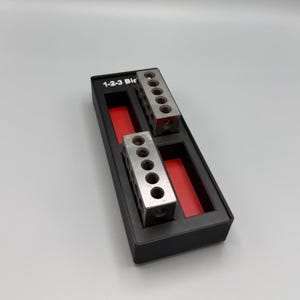 May include: Two rectangular steel 1-2-3 blocks with five circular holes on the top surface, housed in a black plastic storage tray with red interior accents. The tray features the white printed text 1-2-3 Blo on the upper left corner.