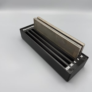 Peut inclure: Un support de pierre à aiguiser noir avec une pierre à aiguiser en métal argenté. Le support comporte trois fentes et le texte "Diamond Sharpening". La pierre est rectangulaire et repose sur le support.
