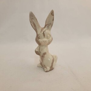 May include: A white ceramic rabbit figurine with brown marbling. The rabbit is standing on all four paws with its ears up and its head tilted to the side.