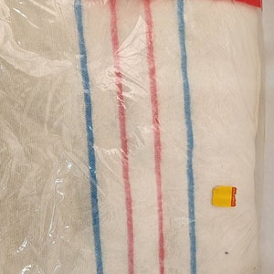 May include: A packaged white towel with blue and red vertical stripes. The towel is wrapped in clear plastic and sealed with a red plastic top. A yellow price tag is visible on the package.