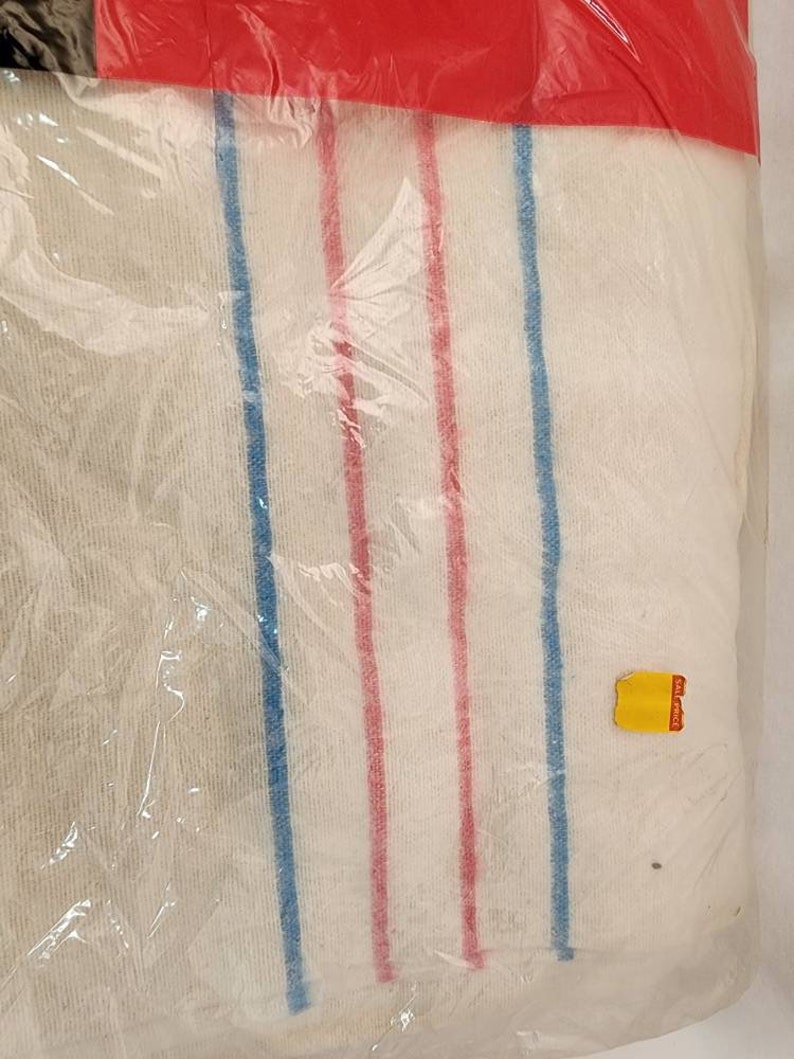 May include: A packaged white towel with blue and red vertical stripes. The towel is wrapped in clear plastic and sealed with a red plastic top. A yellow price tag is visible on the package.