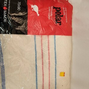 May include: A white sheet-blanket with blue and red stripes, packaged in clear plastic. The packaging includes the text "polar" and "70x90". The blanket is made of flannelette.