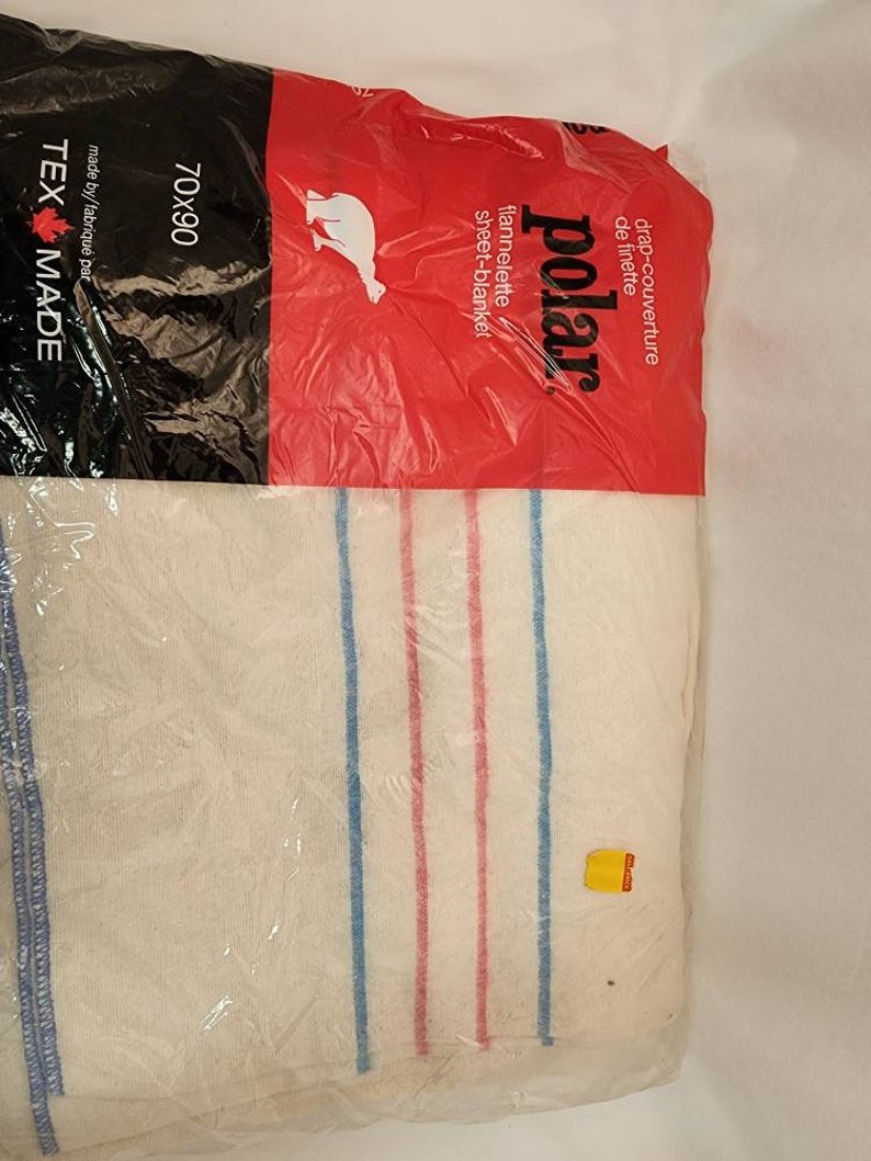 May include: A white sheet-blanket with blue and red stripes, packaged in clear plastic. The packaging includes the text "polar" and "70x90". The blanket is made of flannelette.