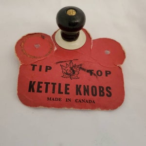 Peut inclure: Étiquette en papier rouge et blanc avec du texte noir qui dit "Tip Top Kettle Knobs Made in Canada". Un bouton noir avec un centre blanc et un dessus en bois est attaché à l'étiquette.