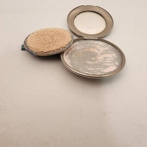 May include: A silver compact with a round mirror and a puff. The compact is open and the puff is sitting on top of it.