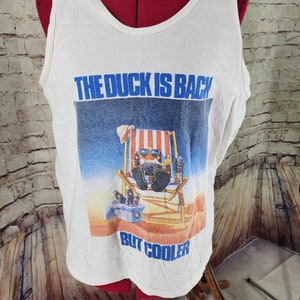 May include: White tank top featuring a blue and white graphic of a cartoon duck wearing sunglasses and relaxing in a beach chair. The text "THE DUCK IS BACK BUT COOLER" is printed on the shirt.