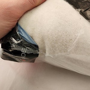 May include: A bundle of off-white knit fabric, partially visible through clear plastic packaging. The fabric appears soft and textured. The packaging has a black label with white text. The item is held by a hand.