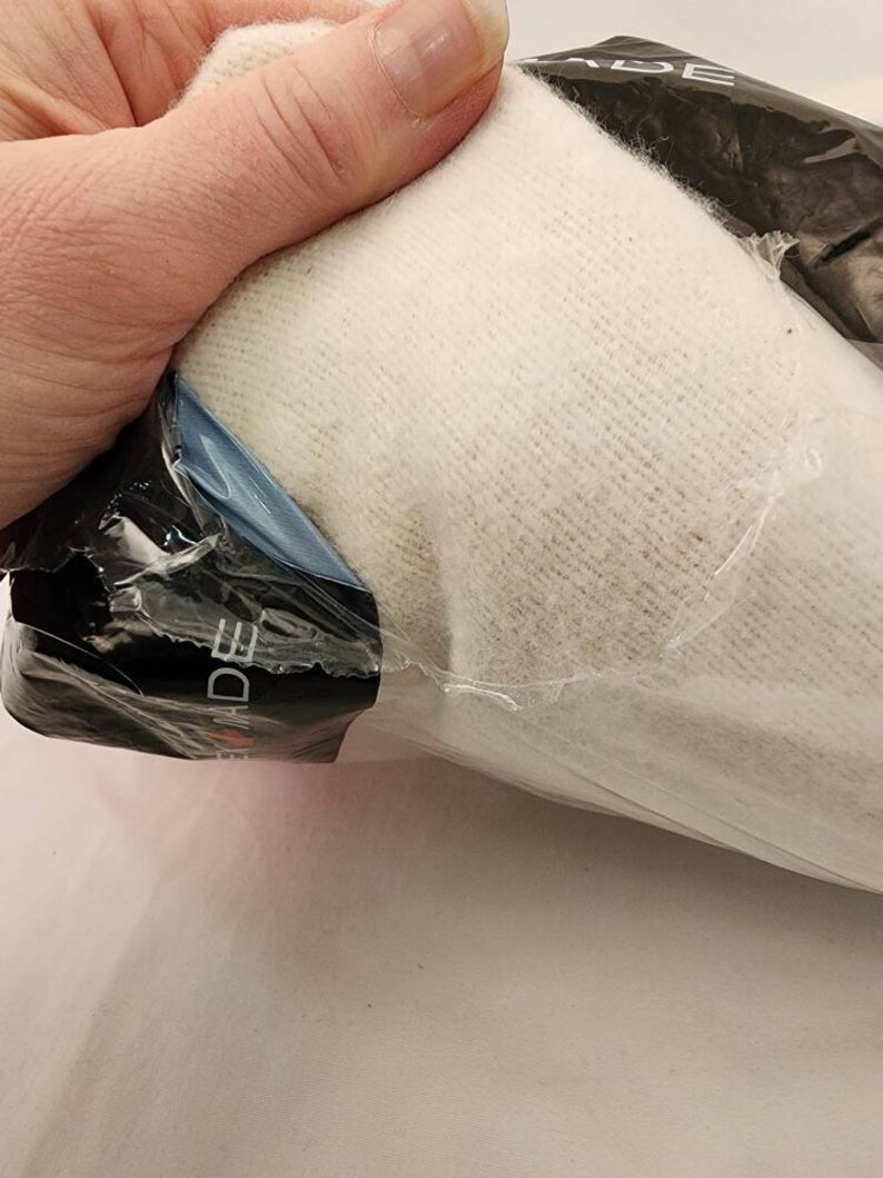 May include: A bundle of off-white knit fabric, partially visible through clear plastic packaging. The fabric appears soft and textured. The packaging has a black label with white text. The item is held by a hand.