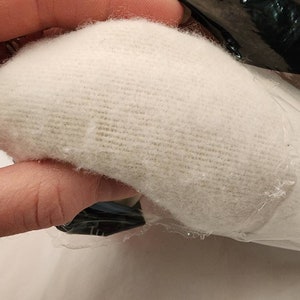 May include: A close-up of a white, textured fabric, possibly a textile or padding material. The fabric is held in a hand, partially visible, and is partially wrapped in clear plastic. The fabric appears soft and fluffy.