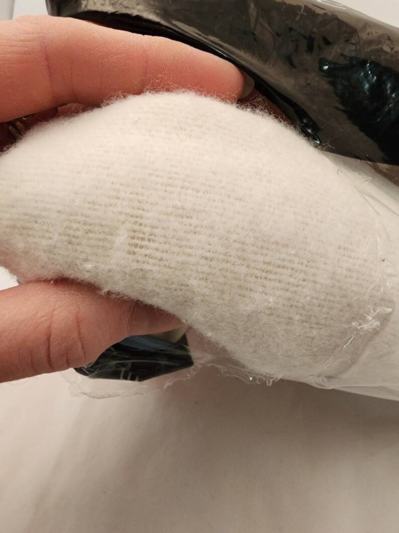 May include: A close-up of a white, textured fabric, possibly a textile or padding material. The fabric is held in a hand, partially visible, and is partially wrapped in clear plastic. The fabric appears soft and fluffy.