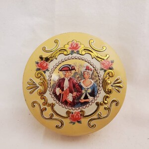 May include: A round, yellow tin box with a gold trim and a floral design. The lid features a couple in 18th-century clothing, surrounded by roses and gold swirls.