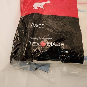 May include: A packaged white and blue striped textile product. The packaging is black with the text "70x90" and "TEX MADE". A polar bear logo is visible on the red top of the packaging.