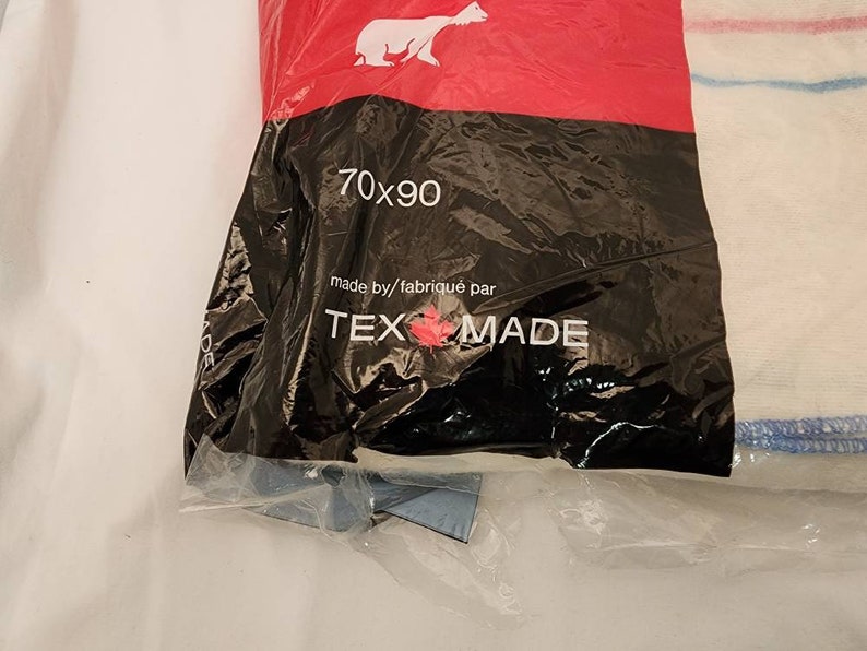 May include: A packaged white and blue striped textile product. The packaging is black with the text "70x90" and "TEX MADE". A polar bear logo is visible on the red top of the packaging.